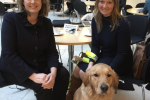 Lana Hempsall sitting in the atrium of Portcullis House with Penny Mordaunt MP and Zorin in his working hguide dog harness