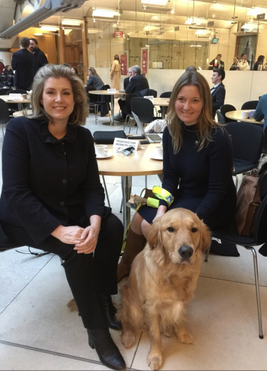 Lana Hempsall sitting in the atrium of Portcullis House with Penny Mordaunt MP and Zorin in his working hguide dog harness